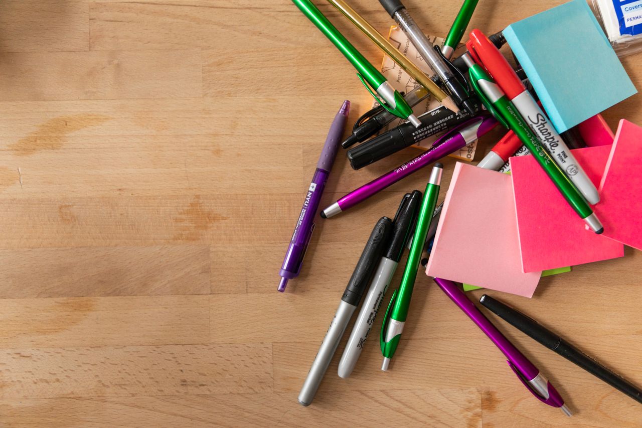 A variety of pens, markers, and sticky notes scattered on a wooden desk.