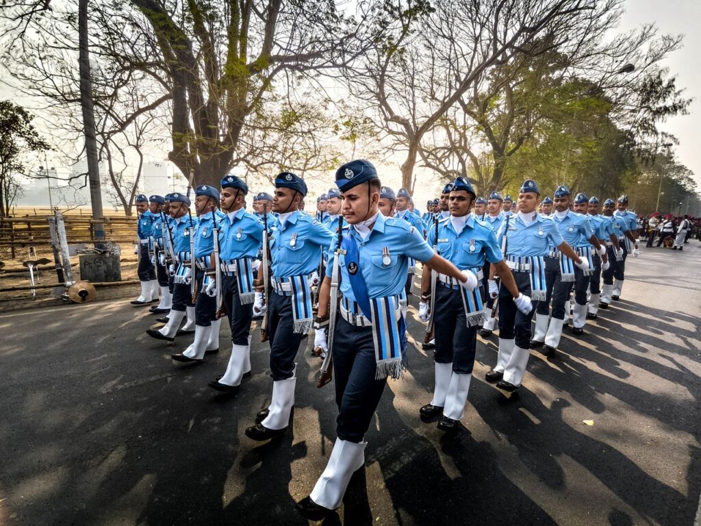A disciplined parade in blue uniforms marches down a sunny road outdoors.